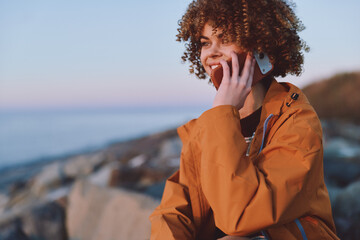 Young woman with curly hair wearing orange jacket smiles while talking on smartphone outdoors near...