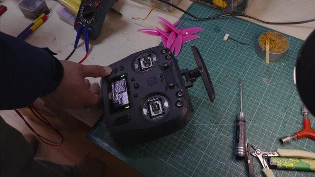 Detailed view of a person adjusting a drone remote controller on cluttered workshop table filled with electronic tools, wires, and model components for engineering and hobby projects