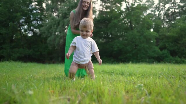 Mother holding son hand walking outdoors. First steps for son with parent support. Family enjoying sunny day. Parent guiding on grass. Family bonds during son first steps. Parent son outdoor adventure