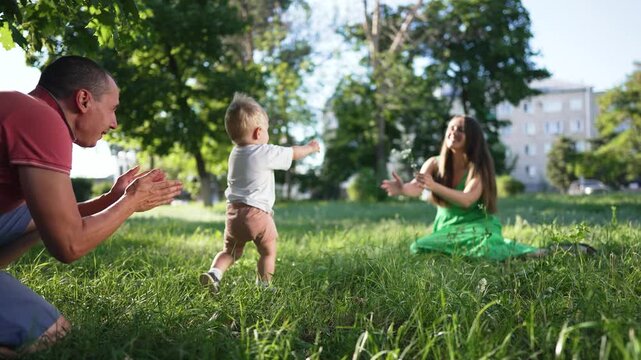 Parents teach son to walk. Father helps son take first steps toward mom in green park. Happy family outdoor bonding. Baby first walk with mom and father. Joyful moments as mom father encourage son.