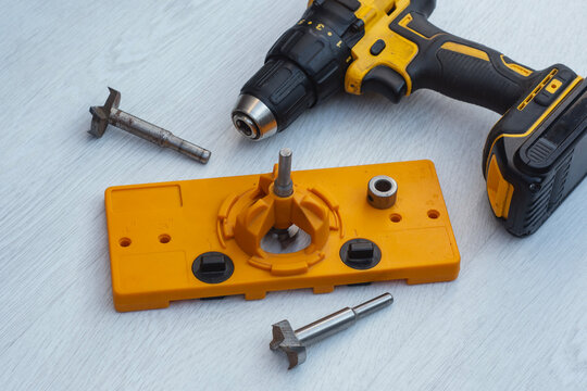 Cordless drill with orange cabinet hinge jig and metal boring bits. Equipment for precision woodworking, furniture assembly and kitchen cabinet hardware installation in home renovation projects.