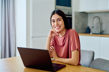 Happy young woman sitting at a table in her modern kitchen, smiling at the camera while using a laptop computer