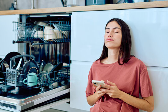 Woman feeling tired and overwhelmed by household chores, leaning against kitchen cabinets, holding a smartphone with eyes closed