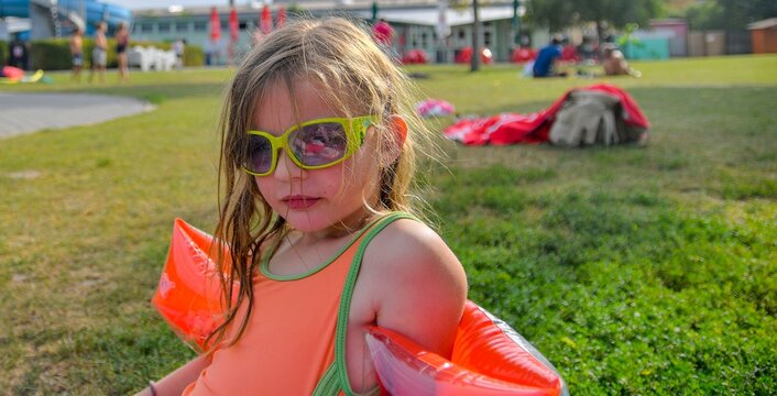 Young girl wearing sunglasses and arm floaties at a water park on a sunny day. A young girl with wet hair and bright sunglasses sits on the grass, wearing ora
