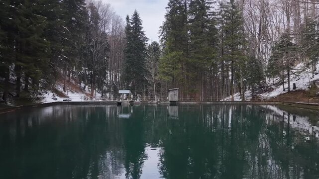 Low altitude drone flight over a small alpine reservoir dam moving toward a forest cabin surrounded by snowy trees in a winter mountain landscape.