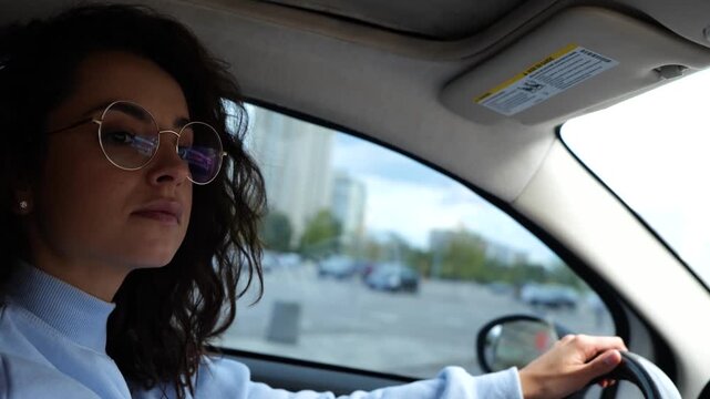 Caucasian young female in stylish glasses with curly hair driving a car in city looking at road. Side view. Close up. Beautiful woman driver beginner practicing driving vehicle