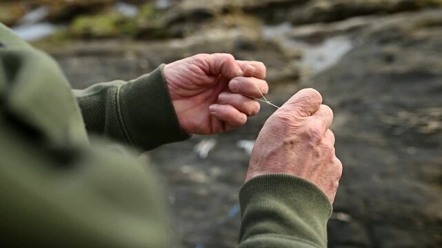 Horizontal close up of fisherman hands tying fishing line and preparing rig for surf fishing on rocky beach, angling knot technique and tackle setup