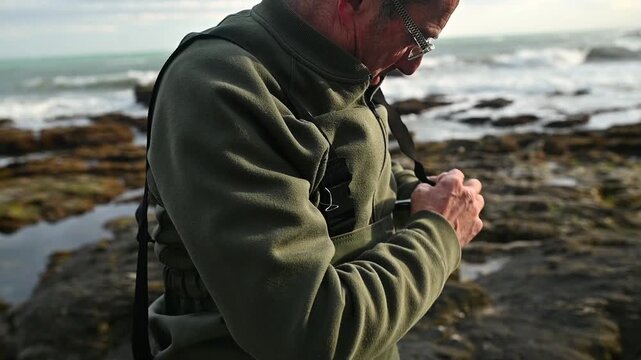 Horizontal medium shot of active senior man adjusting fishing waders vest suspenders on rocky beach, get ready for beach fishing, healthy retirement lifestyle and outdoor wellbeing by the sea
