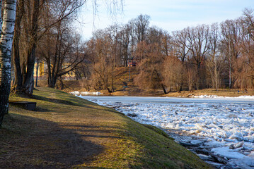 Partly frozen river with drifting ice along a park shoreline, bare trees and a hilltop gazebo under clear winter sunlight. © Aivis