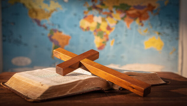 Wooden cross rests upon an open sacred book, set against a blurred background of a world map, symbolizing faith spreading across global cultures and spiritual journey