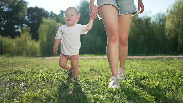 Toddler first steps with mom outdoors. Mom holds toddler hand while learning to walk on green grass. Happy family in nature. Toddler enjoys first steps with mom. Happy Toddler first walking experience