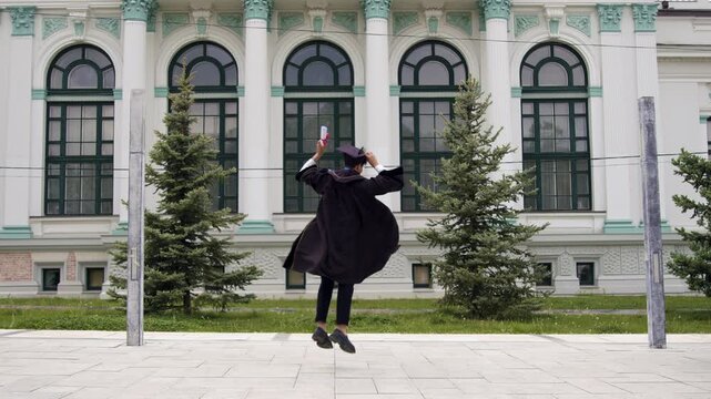 Graduate in black gown and cap jumps joyfully while holding diploma outside a historic building with green trees and large windows in the background
