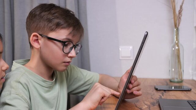 Young boy with glasses intently using a tablet at home, focused on digital learning and e learning content, showcasing modern technology, concentration and study at a table