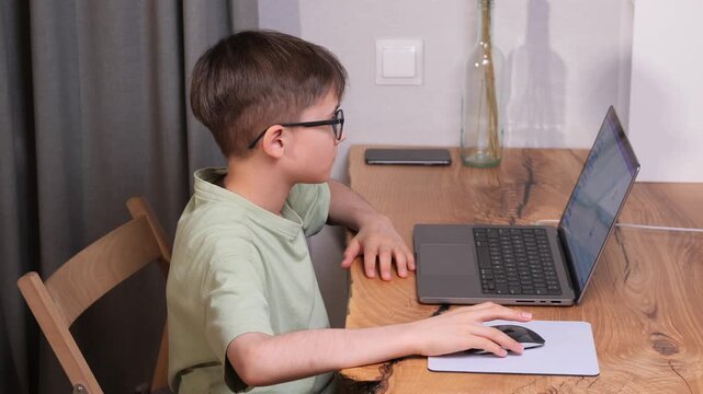 Young male student wearing glasses sitting at a wooden desk, concentrating on his laptop screen while using an external mouse, engaging in remote education and digital learning activities
