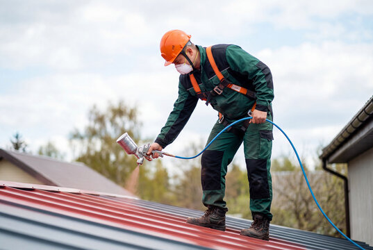 Male worker spray painting red metal roof wearing safety helmet and harness outdoors