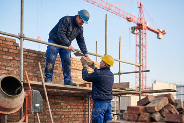 Fototapeta premium Two construction workers on scaffolding laying bricks for a wall with a crane in the background