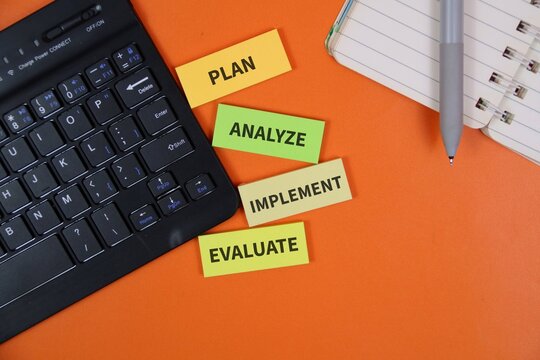 Colorful sticky notes reading Plan, Analyze, Implement and Evaluate arranged beside a keyboard, notebook and pen on an orange desk, illustrating business strategy and planning process.