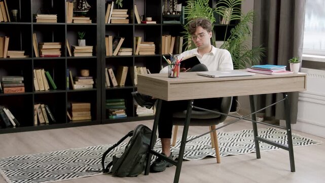 Young male student with curly hair studies at a modern desk surrounded by bookshelves filled with books and a potted plant in a well-lit indoor study space