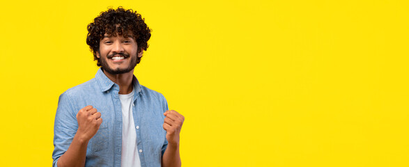 A man with curly hair stands in front of a bright yellow background. He has a big smile and clenched fists, showing excitement and happiness. The scene captures a moment of joy and celebration. © Prostock-studio