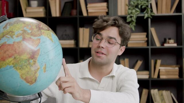 Young male educator with glasses interacts with a globe, pointing to locations while seated at a desk surrounded by bookshelves filled with books