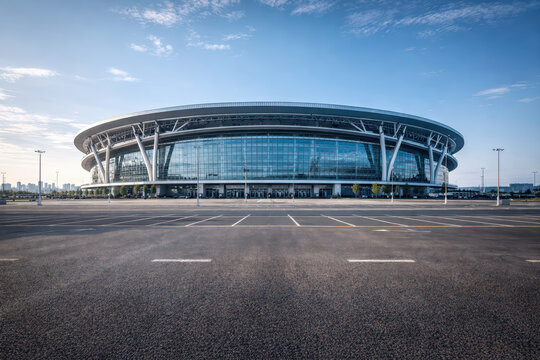 Modern stadium exterior with large glass facade and empty parking lot under blue sky, architectural design with metal supports and urban background