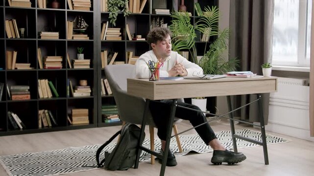 Young male student in white shirt sits at desk in home office, showing signs of stress while surrounded by books and study materials in a cozy environment