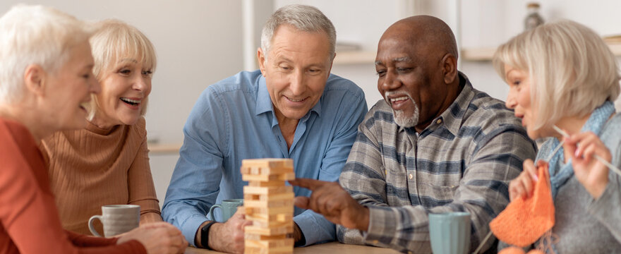 Six seniors are gathered around a table, focused on a game of stacking wooden blocks. They share smiles and laughter while enjoying their time together in a bright indoor space.
