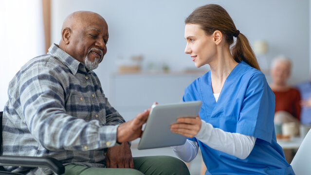 A caregiver helps an elderly man with a tablet while sitting in a clinic. Other patients can be seen in the background. The interaction is friendly and focused on the device.