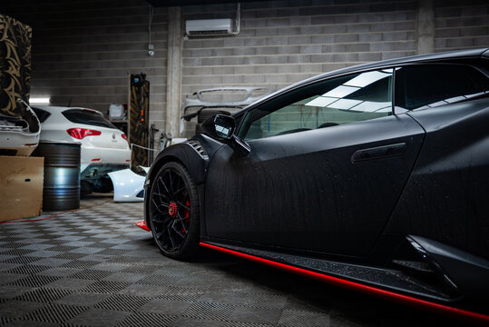 Matte black Lamborghini Huracan shows rear three quarter indoors, red side skirts, center lock wheels, water droplets, oil drums, stacked panels, fluorescent lights.