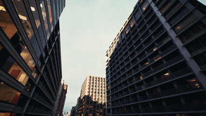 Naklejka premium lowangle skyscraper canyon with warm light, modern facade textures and deep shadow contrasts, repeating window grids and balconies, empty street valley,