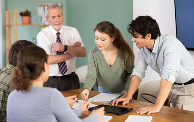 Fototapeta premium Students solve problems together at table in classroom, teacher, standing in background, monitors process