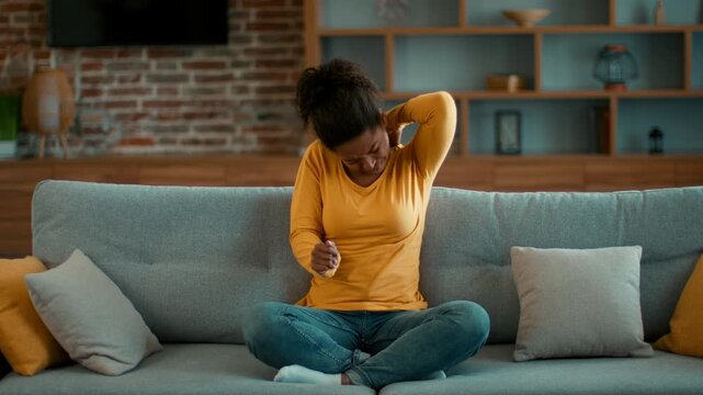 A woman with dark skin is sitting on a couch in a cozy living room while holding her neck. She looks uncomfortable, suggesting she is experiencing neck pain. The space has a modern decor.