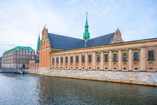 Holmen Church is located in central Copenhagen, Denmark. The building features green spires and brick walls. It sits next to the water under a cloudy sky.