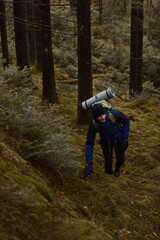 Obraz premium A hiker with a backpack and sleeping mat climbs a mossy, steep hill in a dense evergreen forest. Wearing a blue jacket and beanie, he navigates the wild, overcast woodland during a cold autumn day.
