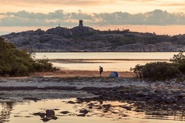 Hiker with backpack standing by a blue tent on a rocky beach at sunset, overlooking a calm bay and a historic coastal tower in the Swedish archipelago. Serene outdoor camping and adventure scene. © Francis