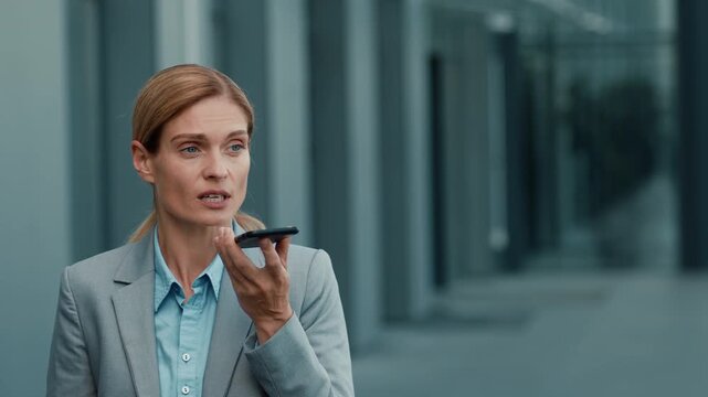 A woman in a business suit is standing outside in an urban area, speaking into her smartphone. She appears to be engaged with technology while surrounded by modern buildings.