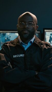 Vertical shot of African American man working as security officer and looking at camera with confident face expression turning away from PC screens with surveilance video feed glowing with blue light