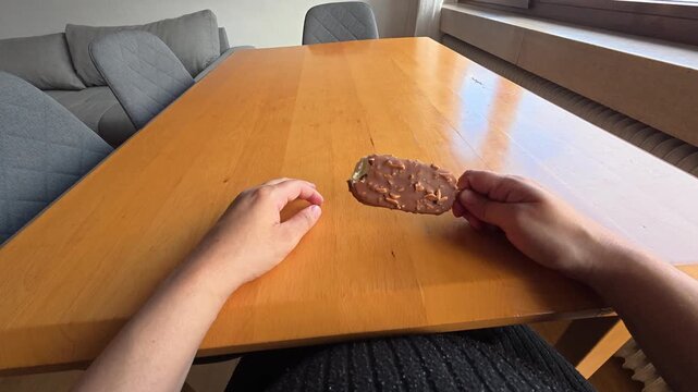 POV of a pregnant woman sitting at a dining table repeatedly biting a chocolate-covered ice cream popsicle with nuts.