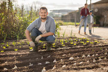 Fototapeta premium Male farmer plants garlic in the garden with his family on the background. Farmer has planted and grows organic seeds.