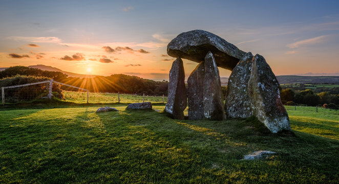 Pentre Ifan Neolithic burial chamber at sunset, surrounded by lush green fields and distant hills under a colorful sky