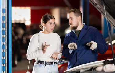 Woman stands near her car in a car service and consults with a male mechanic about the right...