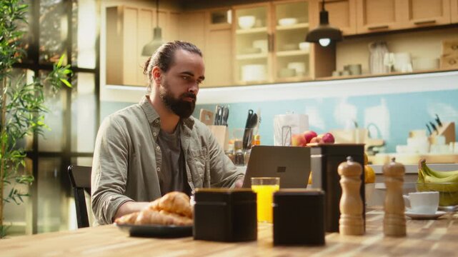 Man working from home in kitchen interior focused on online administration, multitasking using a laptop. Wireless internet and modern technology supports remote work productivity, indoors.