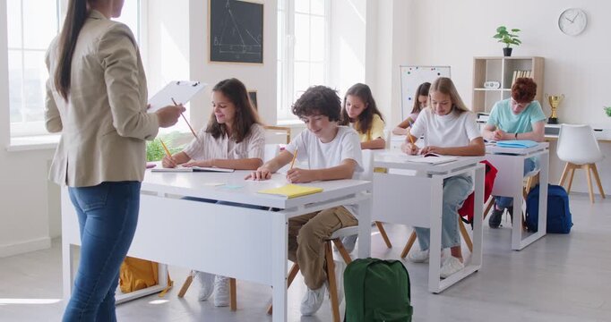 In school classroom, teacher conducts lesson for group of children sitting at white desks. Each student is focused on his work, writing notes under teacher dictation. Concept of modern education.