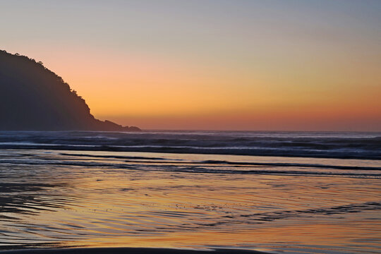 Golden sunset on the beach,  small waves breaking on wet  sand, hill still in darkness, light relflection on sky and water. Bertioga city, Brazil