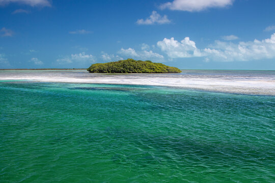 A small tropical island surrounded by green water and blue sky with clouds