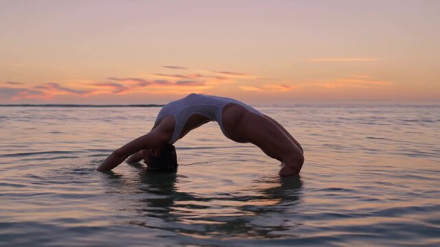 Flexible woman practicing yoga pose in the ocean at sunset