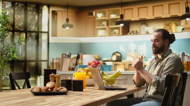 Man cracking knuckles, nervous habit at home, repetitive involuntary popping sound, person focused on tasks and using laptop in the kitchen for teleworking and multitasking at the wooden table.