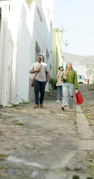 Vertical video: Diverse couple walking downhill cobblestone lane carrying coffee teenager following