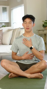 Vertical video: Asian man after closing eyes inhaling and exhaling, meditating on yoga mat at home
