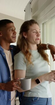 Vertical video: Diverse couple holding mugs by sliding glass door smiling, curtains filtering light